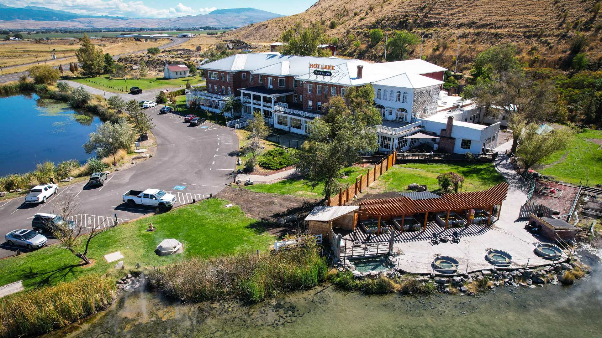 Aerial summer view of Hot Lake Springs Resort, showing soaking tubs, the soaking area pergola, Thermal Pub & Eatery deck, front parking, lodge building, clear Hot Lake in foreground, and Blue Mountains in the distance.