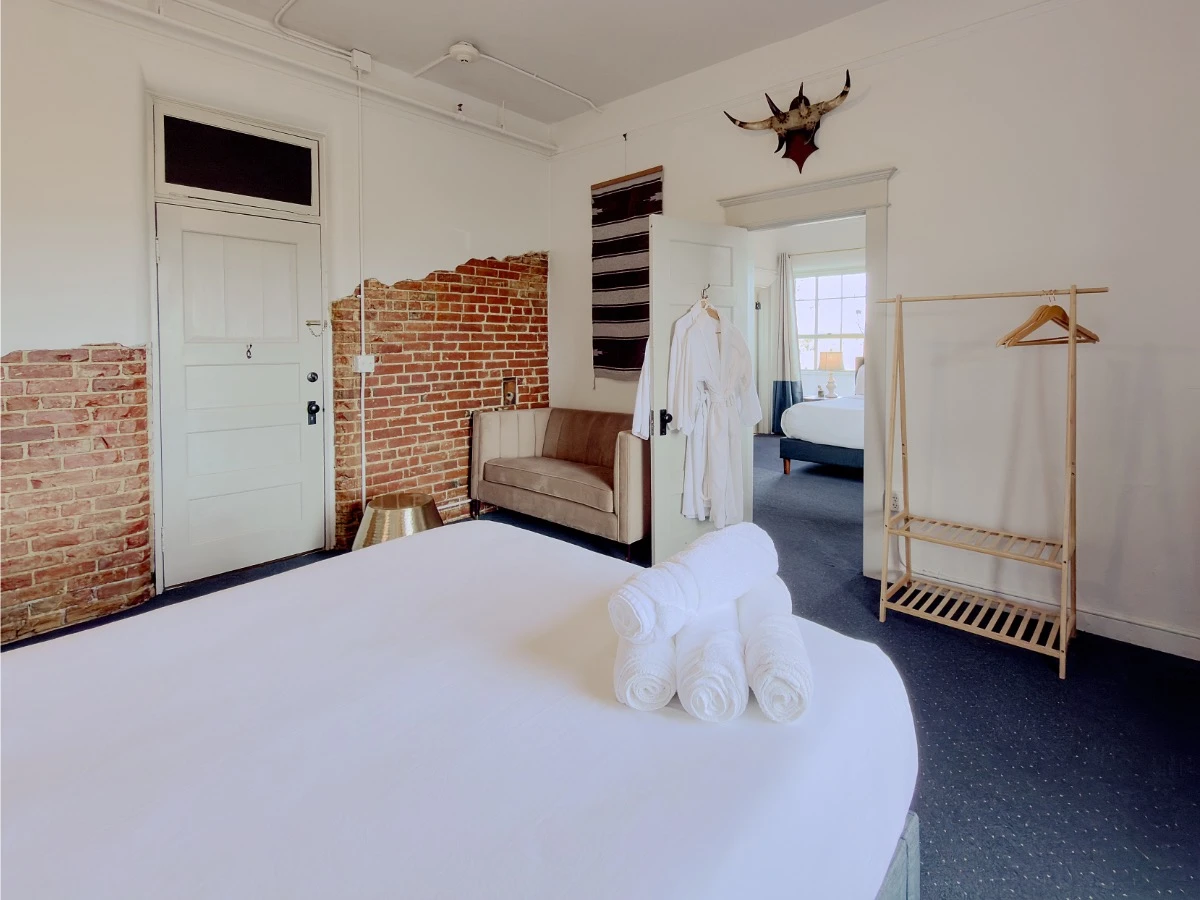 View across foot of bed in Room 113 at Hot Lake Springs Resort into second room, with small loveseat and exposed brick wall on left.