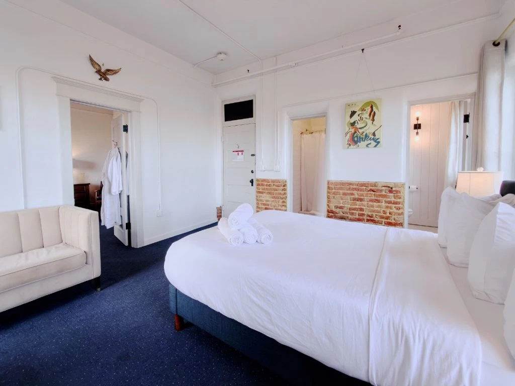 Bed in foreground of Room 113 at Hot Lake Springs Resort, doorway to second bedroom on left and bathroom door on right, shower door in center, with half exposed red brick wall and open grey curtains.