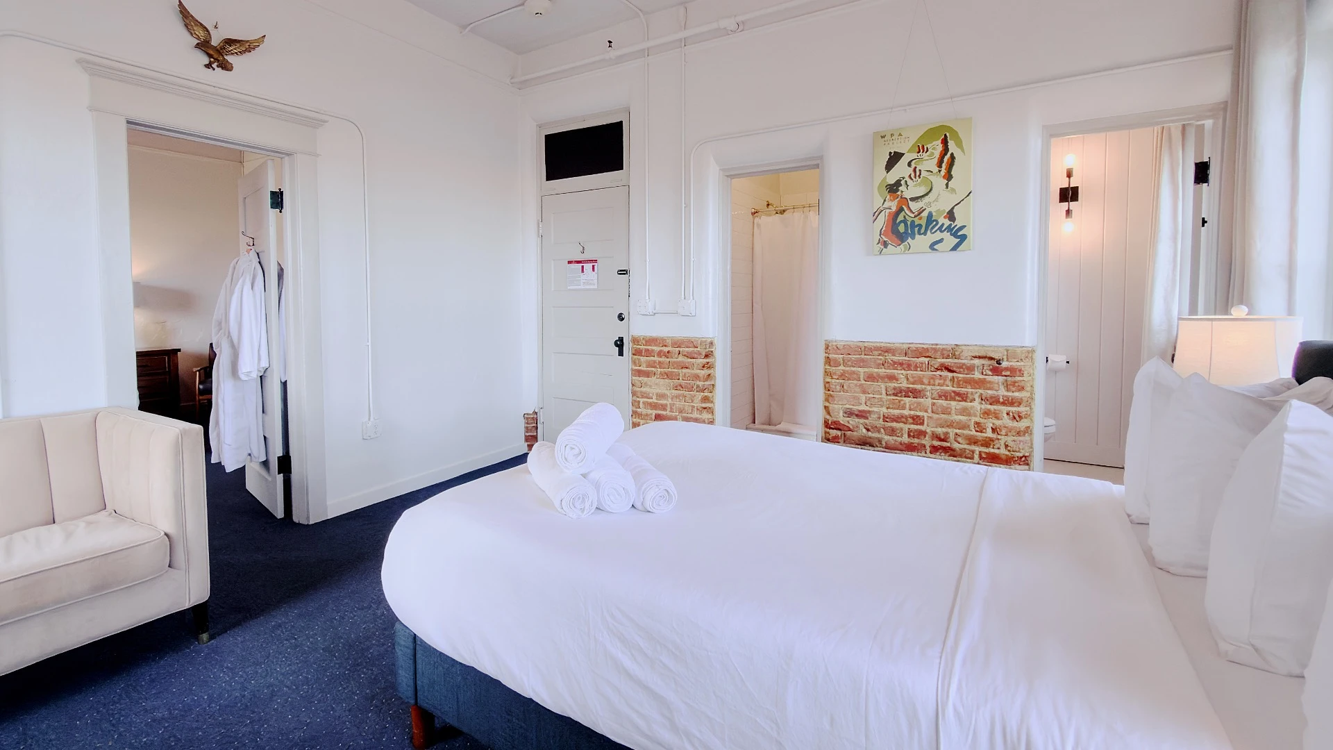 Bed in foreground of Room 113 at Hot Lake Springs Resort, doorway to second bedroom on left and bathroom door on right, shower door in center, with half exposed red brick wall and open grey curtains.