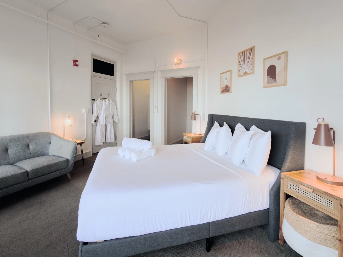 View across bed in Room 115 at Hot Lake Springs Resort with nightstands and lit lamps, entry, closet and hall visible, partial grey sofa on left.