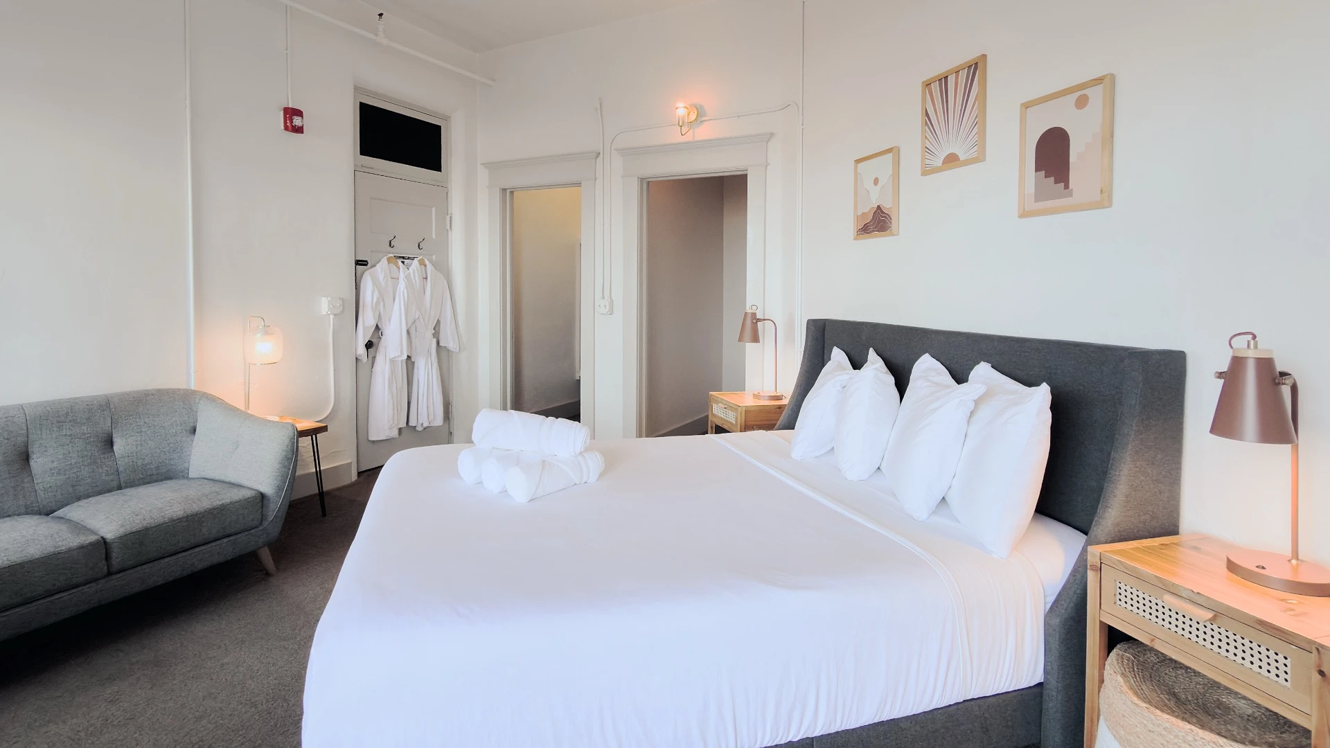 View across bed in Room 115 at Hot Lake Springs Resort with nightstands and lit lamps, entry, closet and hall visible, partial grey sofa on left.