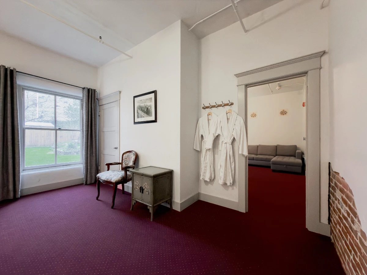 View from bedroom in Room 118 at Hot Lake Springs Resort looking toward living room, doorway on right with partial couch visible, bathrobes hanging on wall, single chair and end table, bathroom entry and window on left showing courtyard.