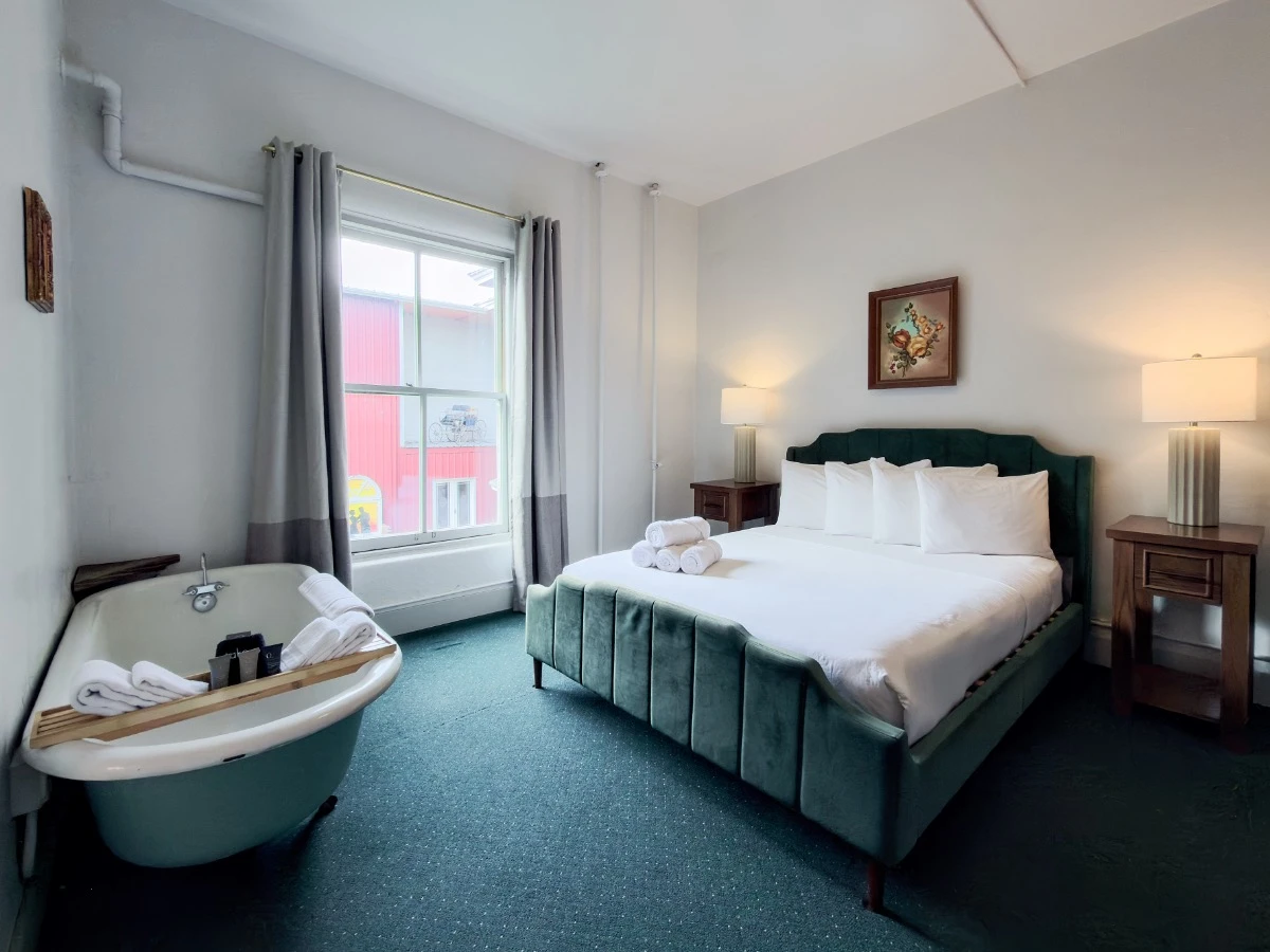 Bed with lit lamps on nightstands in Room 202 at Hot Lake Springs Resort, window on left, clawfoot soaking tub visible near foot of bed against wall.