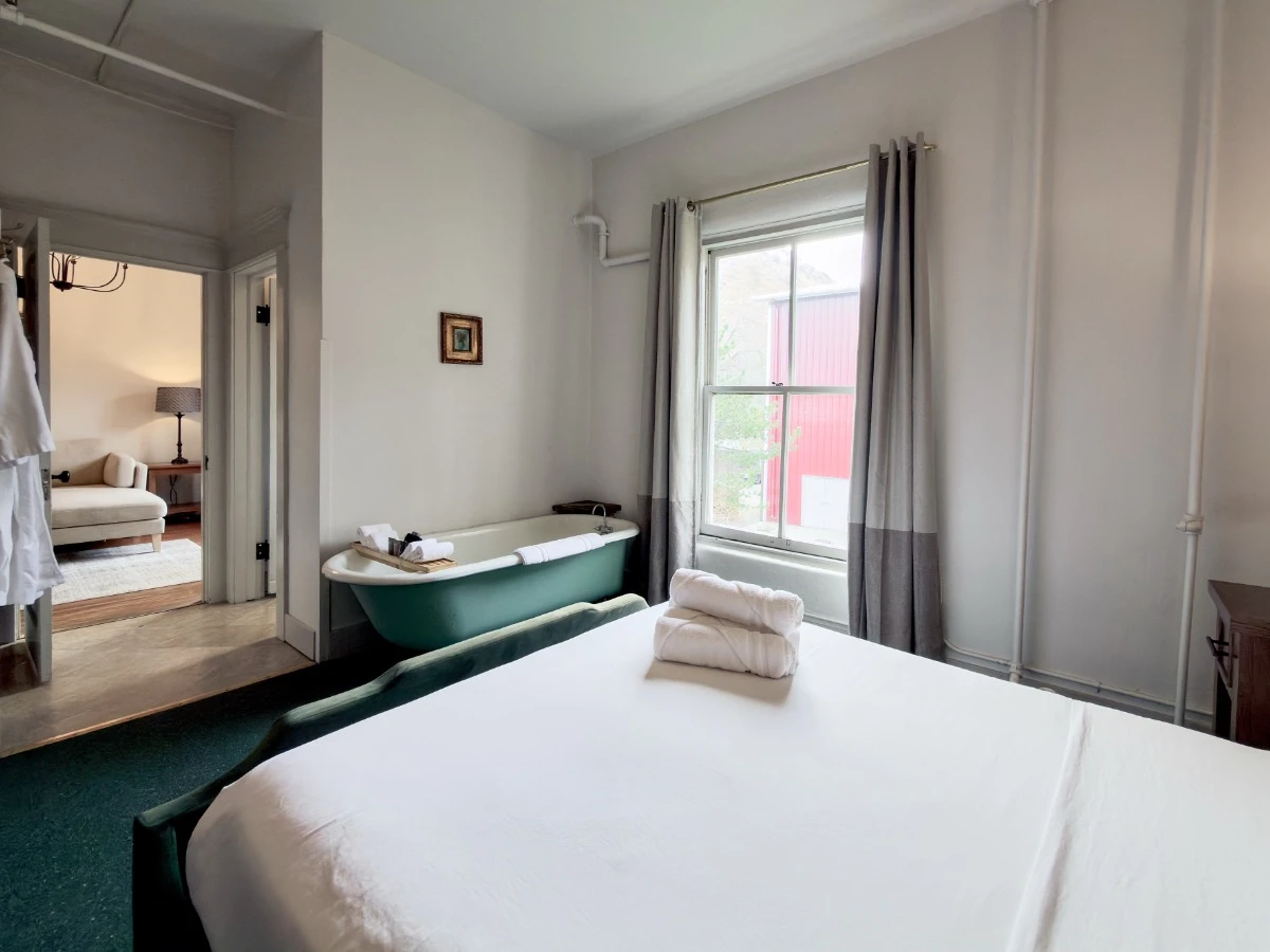 View across foot of bed in Room 202 at Hot Lake Springs Resort, clawfoot soaking tub visible, window on right, entry to living room with couch on left.