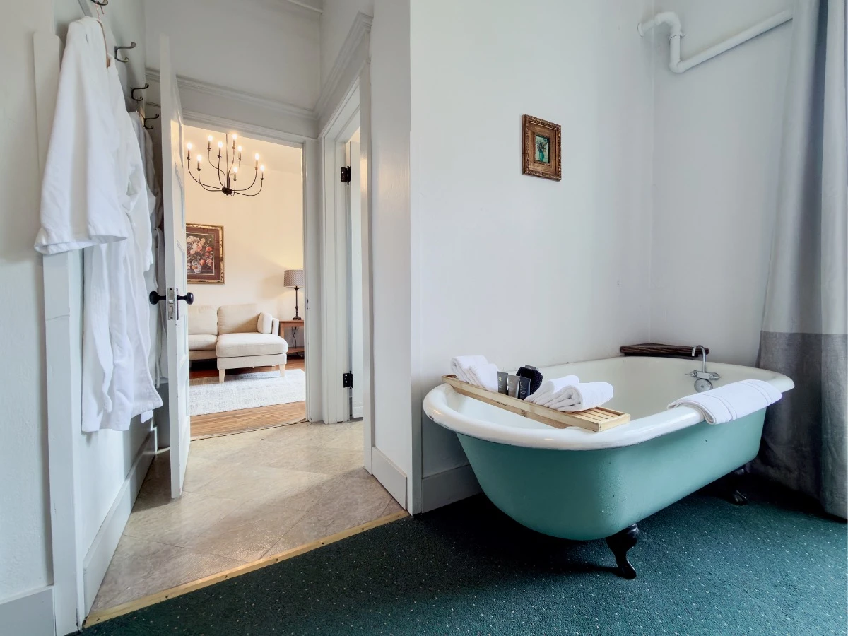 In bedroom of Room 202 at Hot Lake Springs Resort looking back toward living room, clawfoot soaking tub on right, robes hanging and entry to living room with couch visible in background.
