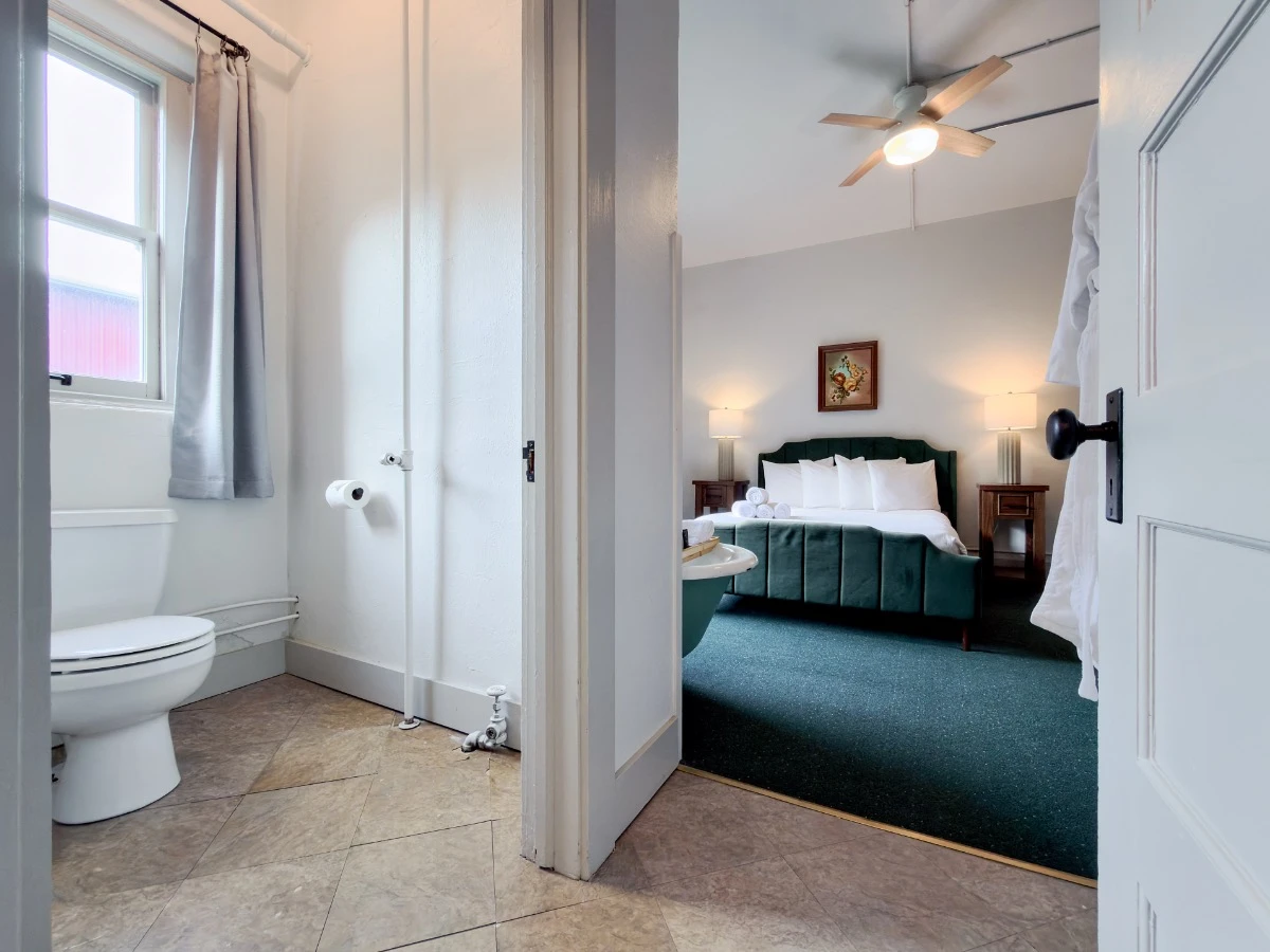 View from living room doorway in Room 202 at Hot Lake Springs Resort looking toward bedroom, bathroom toilet with window on left, bed partially visible on right.