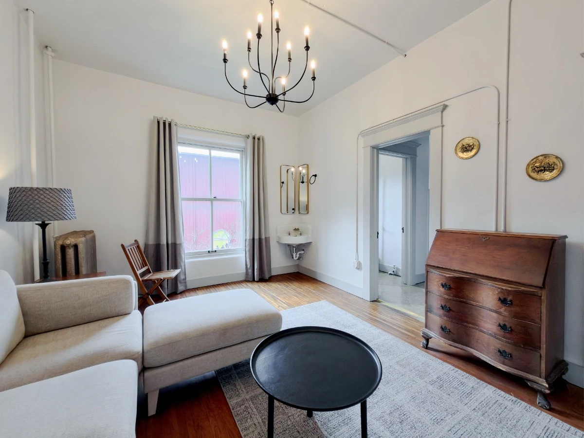 View across living room in Room 202 at Hot Lake Springs Resort, window on left, corner handwashing sink with mirror in center, doorway to bedroom and bathroom on right, partial couch on left, wood 3-drawer unit with fold-down writing desk on right.