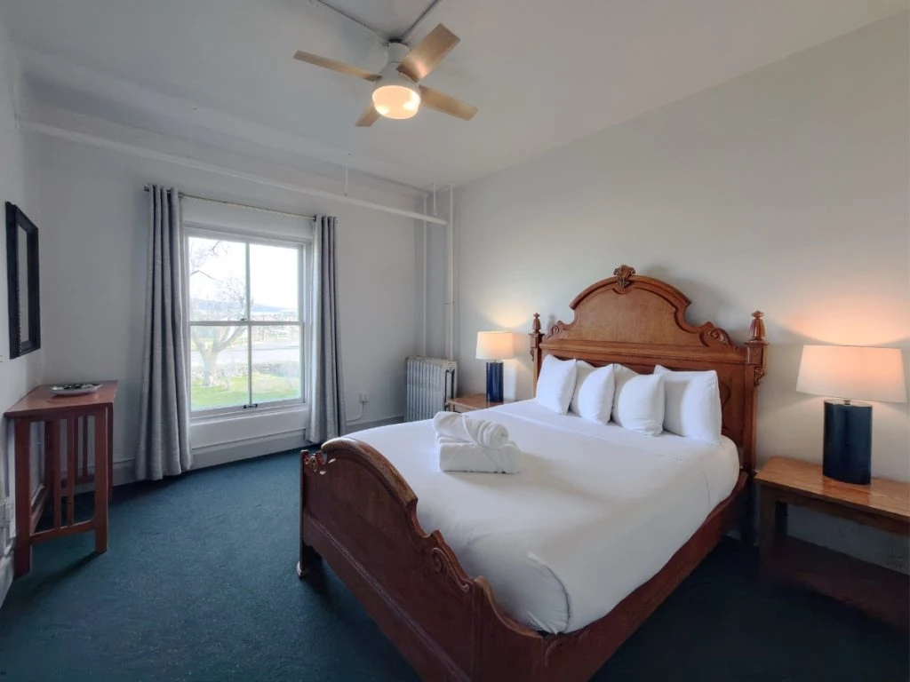 Bed with lit lamps on wood nightstands in Room 203 at Hot Lake Springs Resort, window across from bed, wood standup table/desk on left, blue/green carpet and open grey curtains.