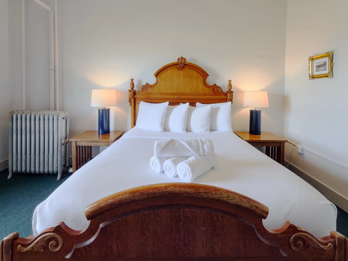 View across foot of bed in Room 203 at Hot Lake Springs Resort looking at head of bed with wood headboard and footboard, nightstands on each side.