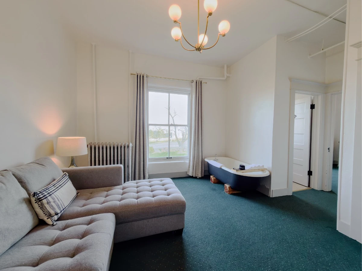Living room in Room 203 at Hot Lake Springs Resort with light gray sofa and lounge section on left, pillow on couch, end table with lit lamp, window in center background, clawfoot soaking tub just right of window, open door to bathroom on right.