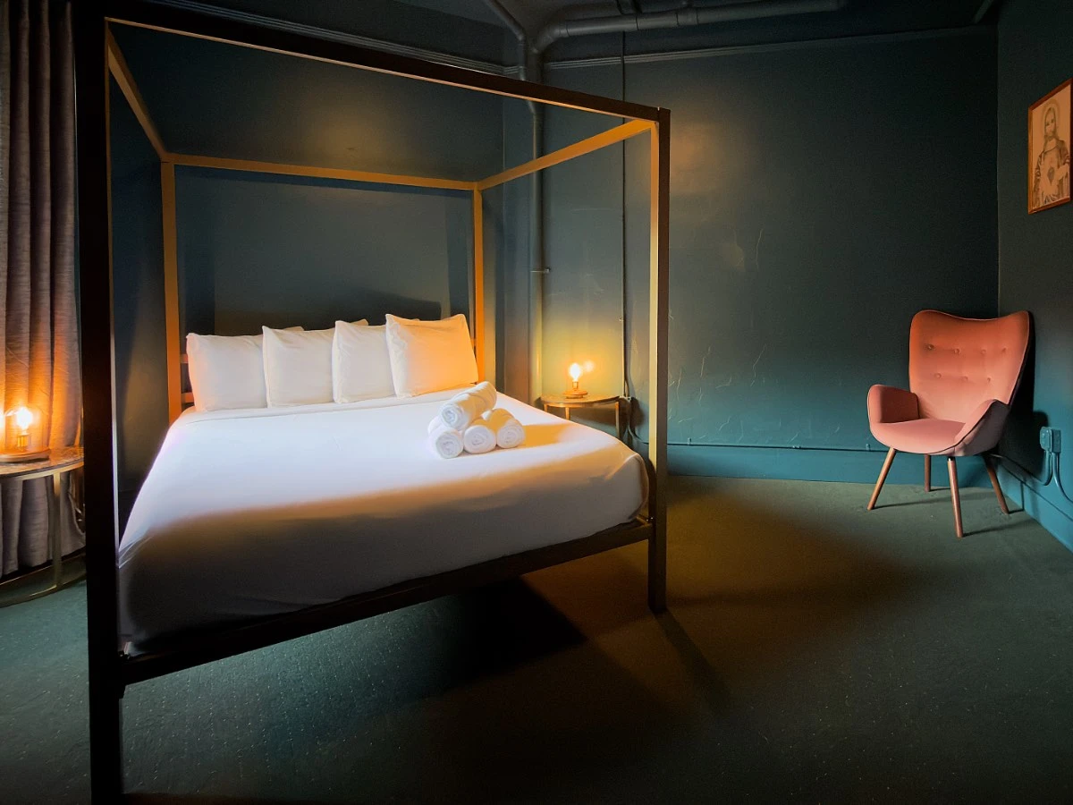 Bedroom in Room 206 at Hot Lake Springs Resort with bed on left, nightstands and lit lamps on each side, orange chair in right back corner, dark green carpet and dark blue/green walls.