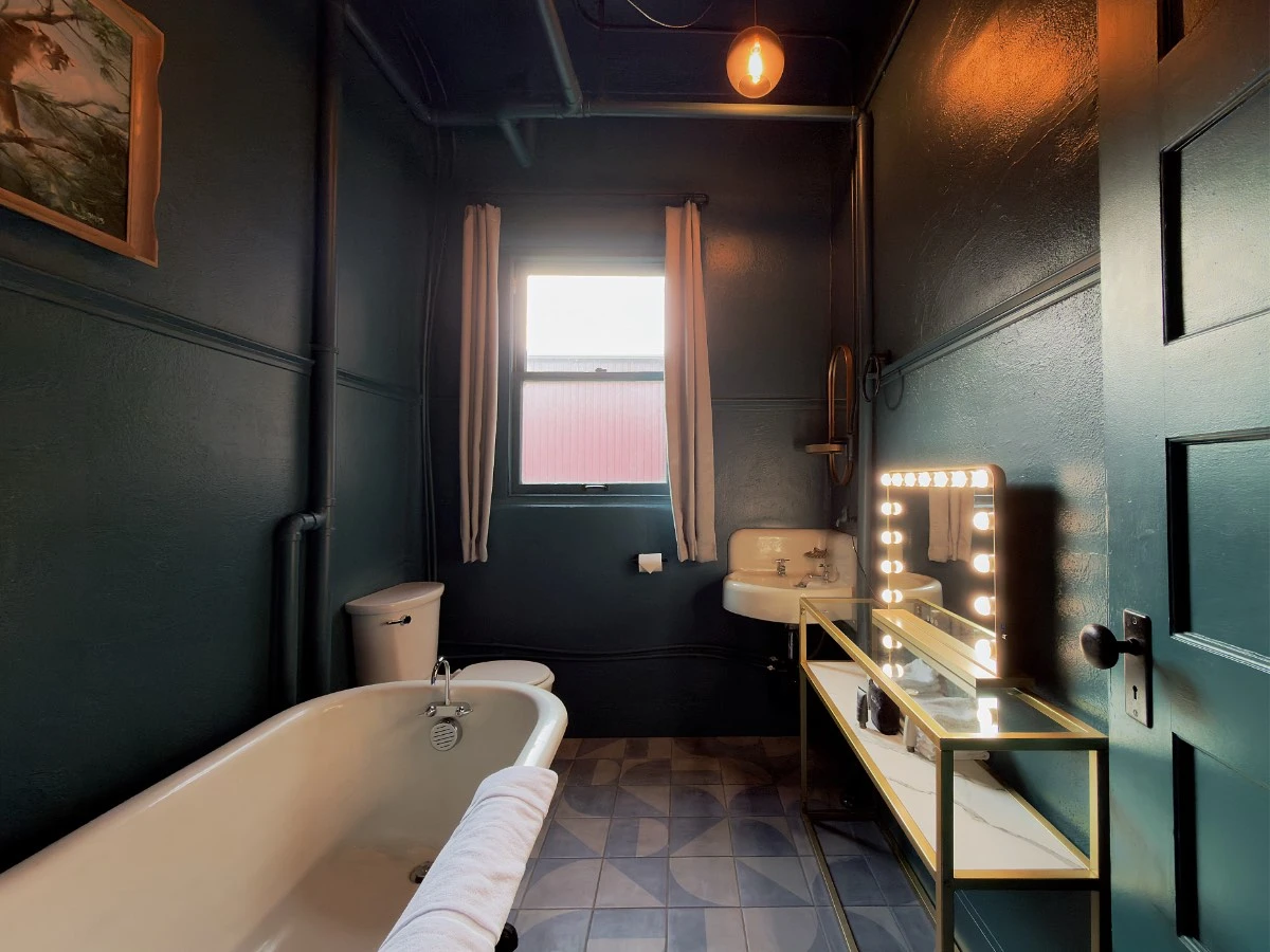 Bathroom in Room 206 at Hot Lake Springs Resort with clawfoot soaking tub on left, toilet at far end, hand sink and table with mirror on right, window on center back wall.