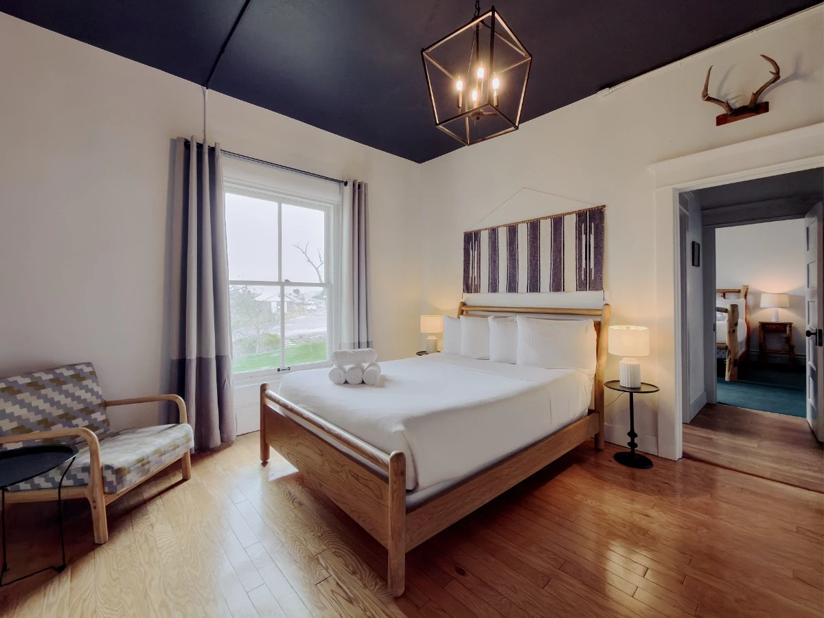 Bed with wood frame in Room 207 at Hot Lake Springs Resort, nightstands with lit lamps on each side, window across from bed, single chair on left, doorway to second bedroom on right.