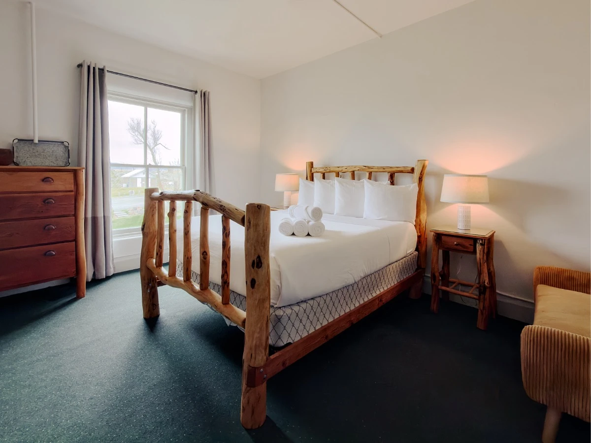 Further back view of wood timber bed in Room 207 at Hot Lake Springs Resort with dark green carpet, nightstands and lit lamps on each side, partial chair on right, wood dresser on left.