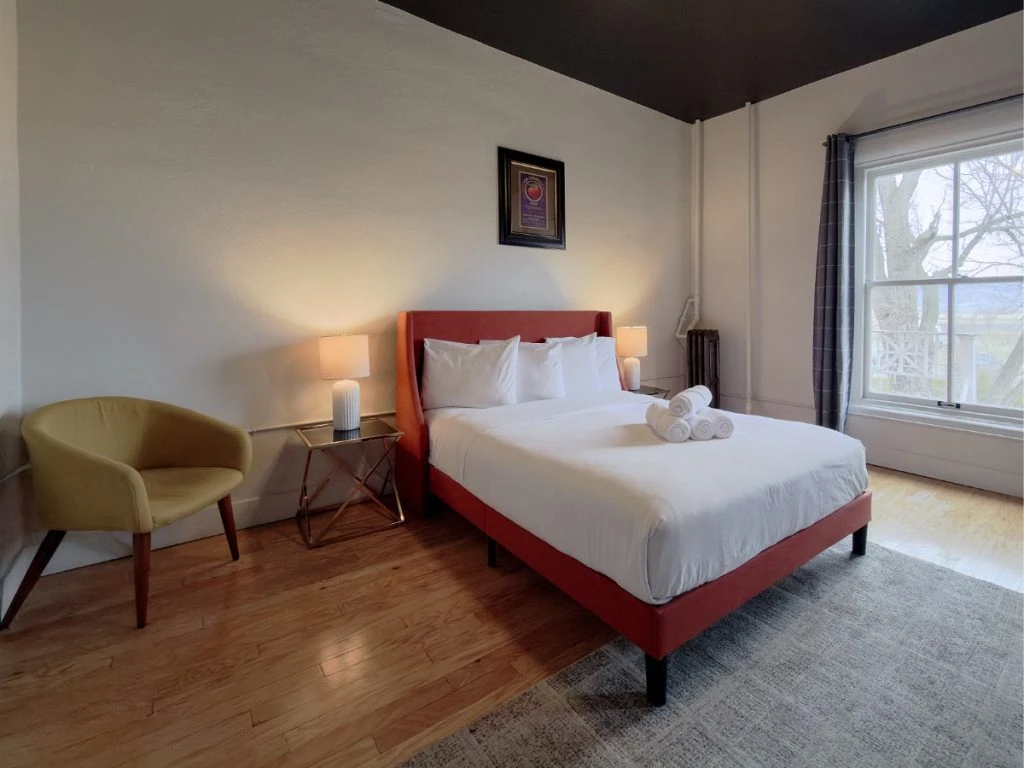 Bed with red/orange frame and headboard in Room 207 at Hot Lake Springs Resort, yellow chair on left, nightstands with lit lamps on each side, window on right, grey rug at foot of bed.