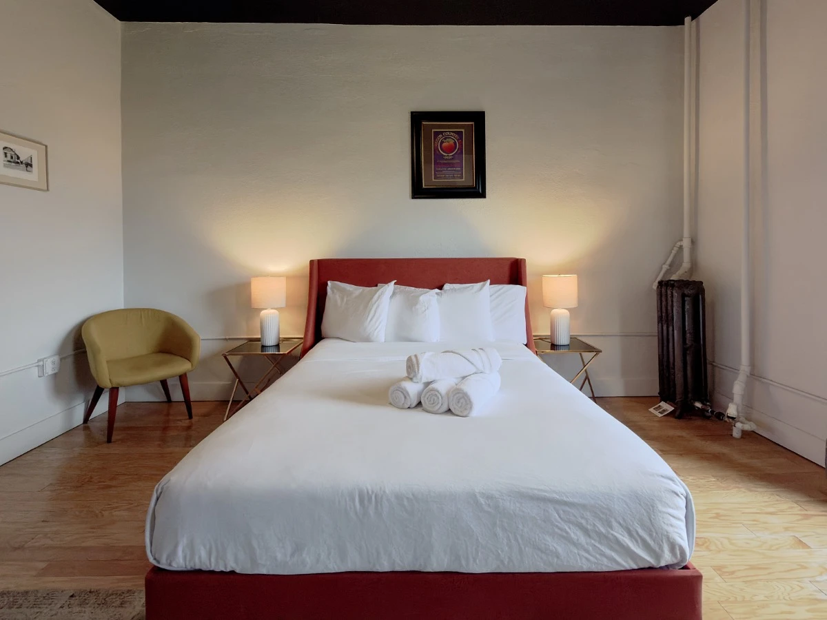 View from foot of bed in Room 207 at Hot Lake Springs Resort looking toward head of bed with red/orange frame, nightstands on each side, yellow chair on left.