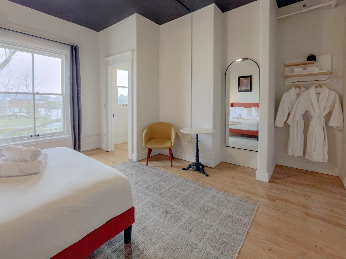 Partial view of bed and window in Room 207 at Hot Lake Springs Resort, closet door with window on far side, yellow chair and round bistro table with white top on right, mirror reflecting bed, bath robes to the right of mirror.