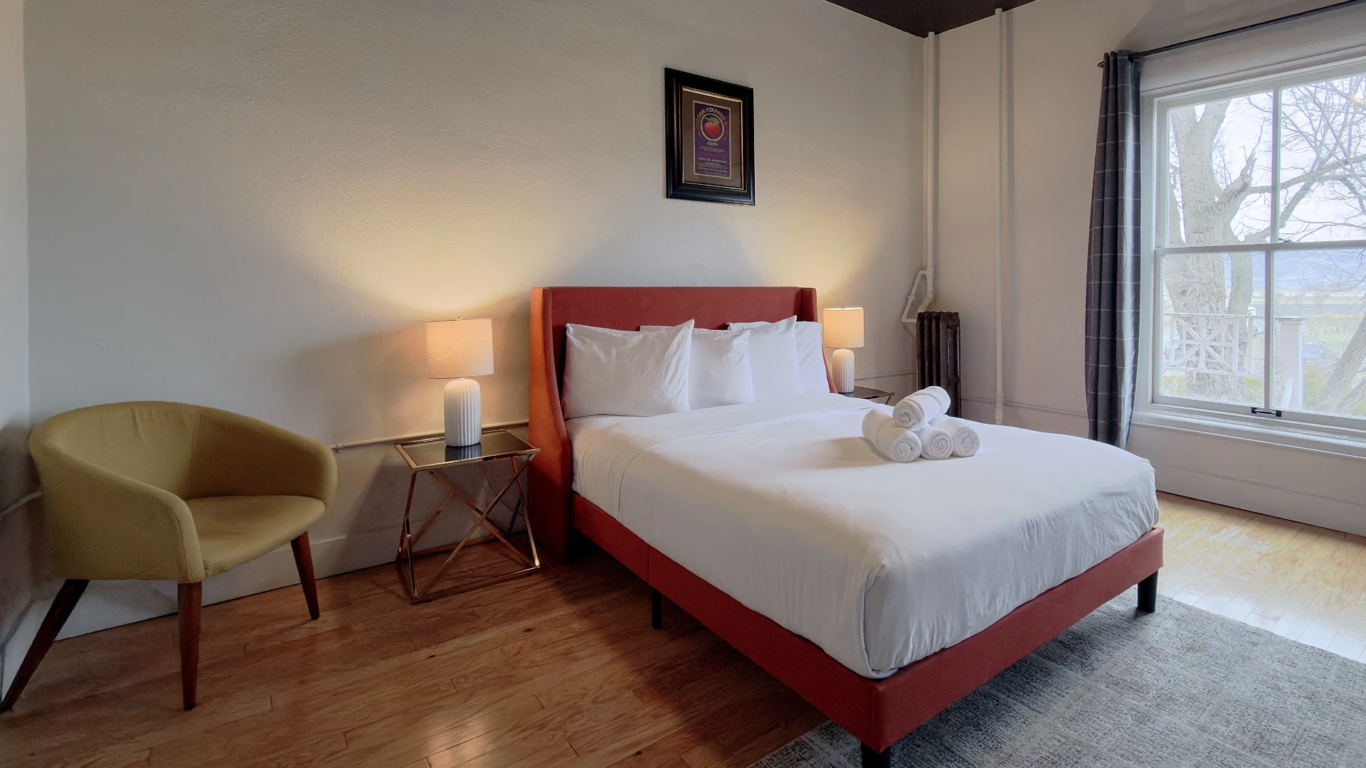 Bed with red/orange frame and headboard in Room 207 at Hot Lake Springs Resort, yellow chair on left, nightstands with lit lamps on each side, window on right, grey rug at foot of bed.