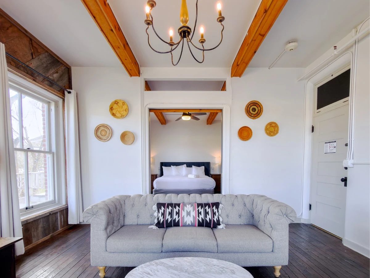 Well-lit historic lodge room at Hot Lake Springs Resort with grey sofa, marble coffee table, dark wood floors, rustic timber beams, and bed visible through open sliding doors.