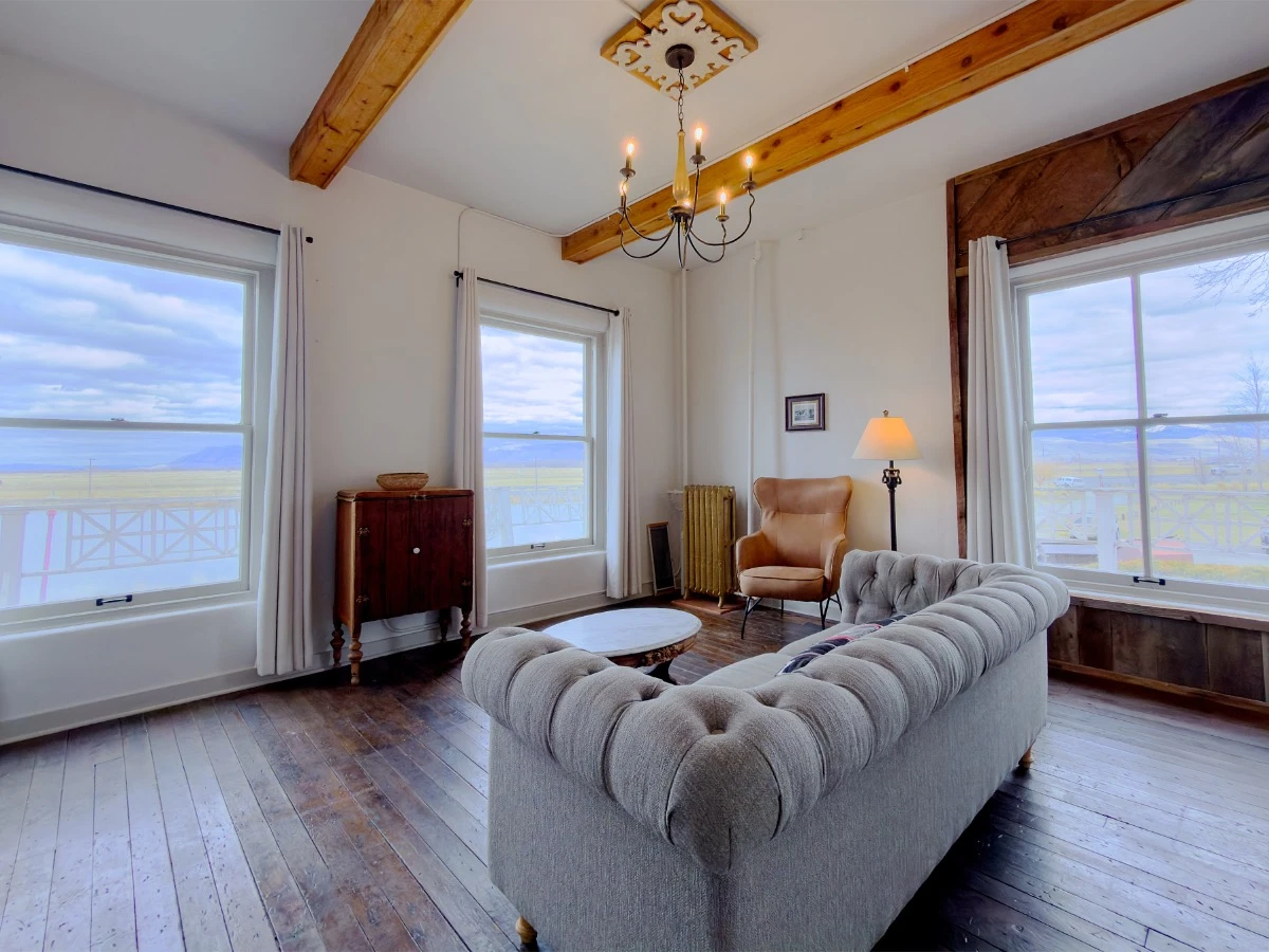 Living room in Room 213 at Hot Lake Springs Resort with grey couch in center, three windows showing views and natural light, cupboard and single chair in background between windows.