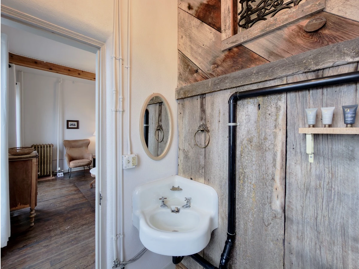 Bathroom in Room 213 at Hot Lake Springs Resort looking at small corner sink with mirror above, rustic wood walls, view out door into living room.