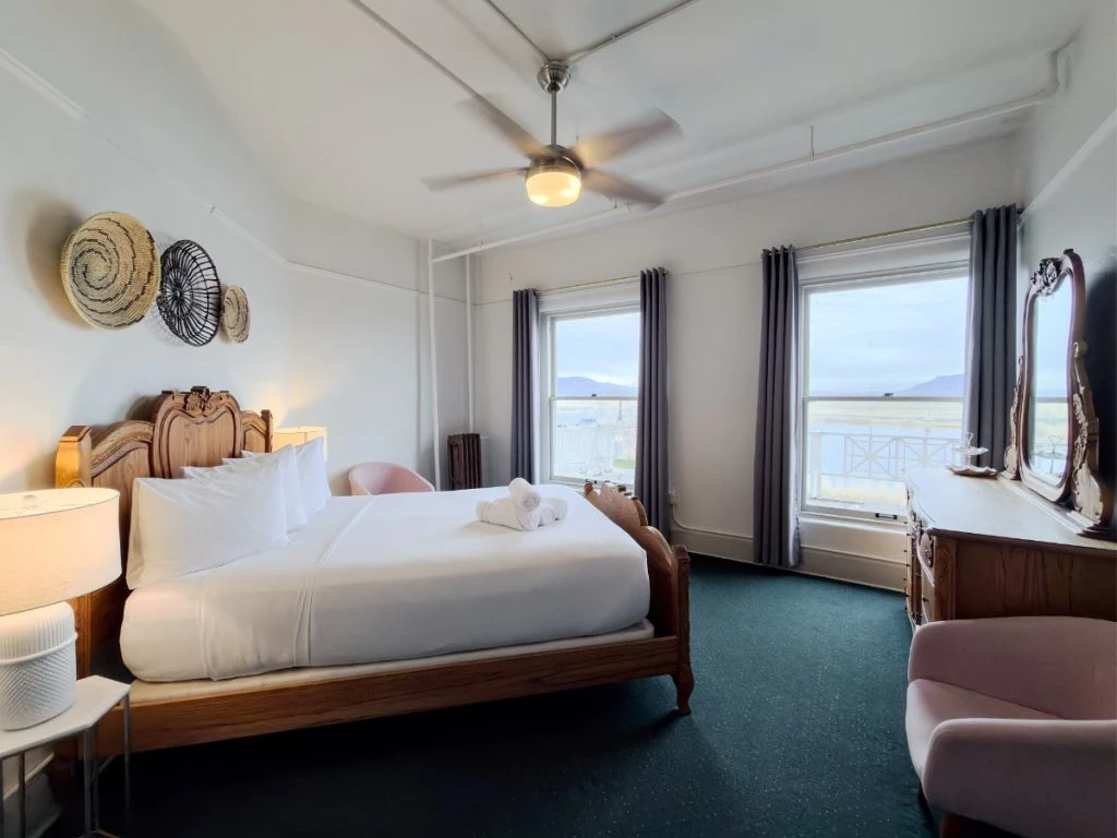 Bed with elegant wood frame in Room 213 at Hot Lake Springs Resort, nightstands with lit lamps on each side, two windows with valley views across from bed, dresser with mirror and chair on right.