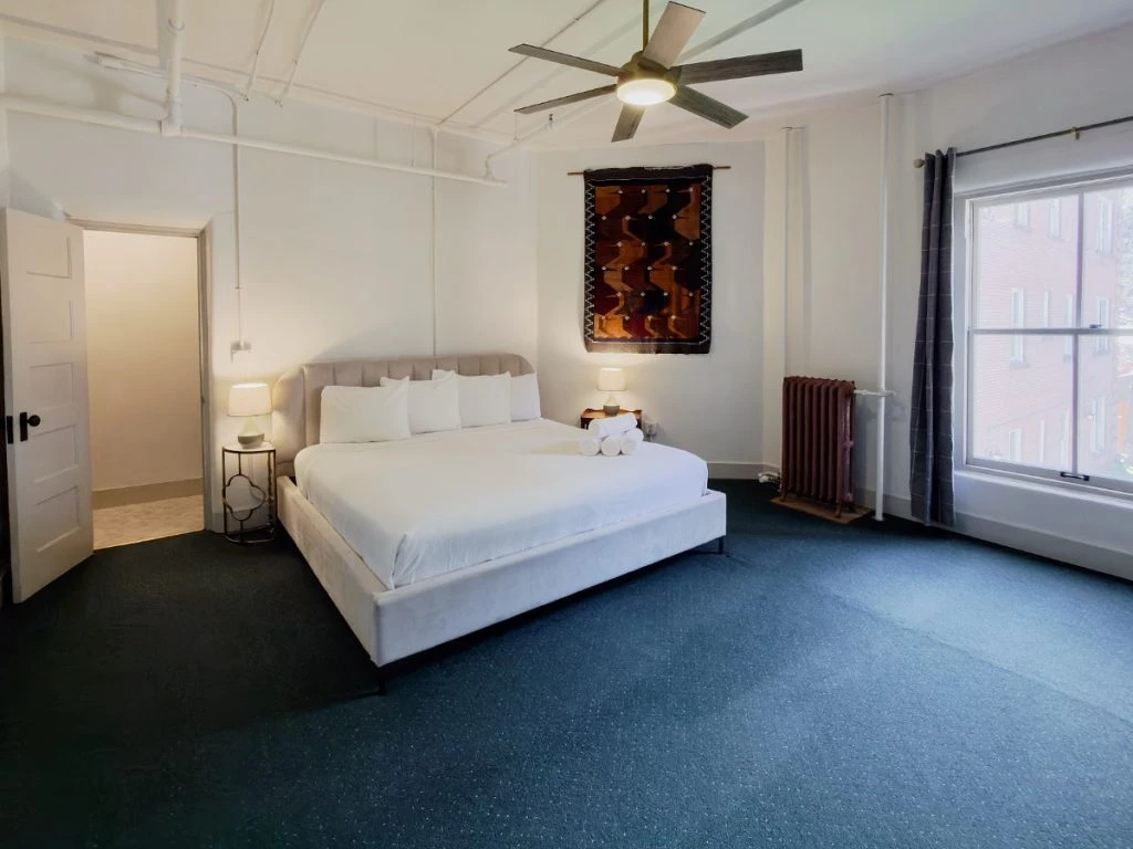 Bed with ceiling fan above in Room 218 at Hot Lake Springs Resort, open door to shower on left, window on right looking toward courtyard, dark blue/green carpet and open dark grey curtains.