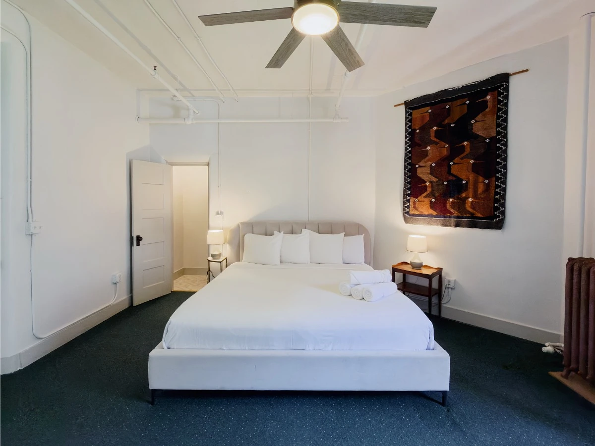 Straight view at foot of bed in Room 218 at Hot Lake Springs Resort looking toward headboard, open shower room door on left, natural light mixing with lit lamps, ceiling fan above bed.
