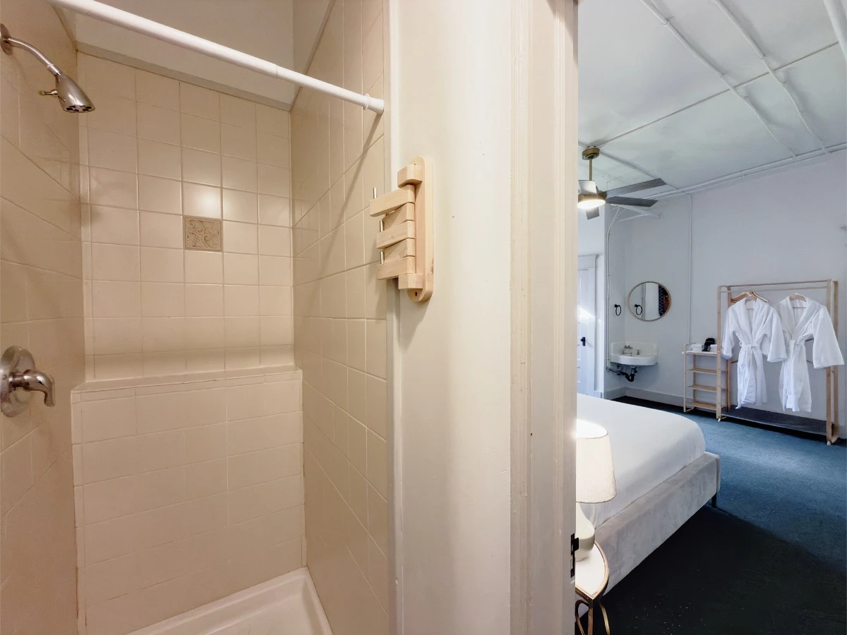 Tile shower in Room 218 at Hot Lake Springs Resort on left, view out shower room door into bedroom with partial bed, hand sink and bath robes in background on right.