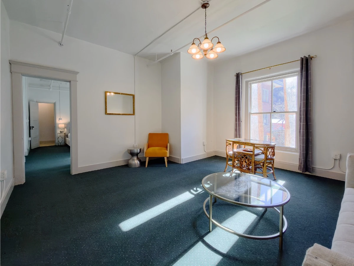Living room in Room 218 at Hot Lake Springs Resort looking toward bedroom doorway, orange chair on far side with mirror on wall, oval table with chairs by window, partial sofa and circle coffee table on right.