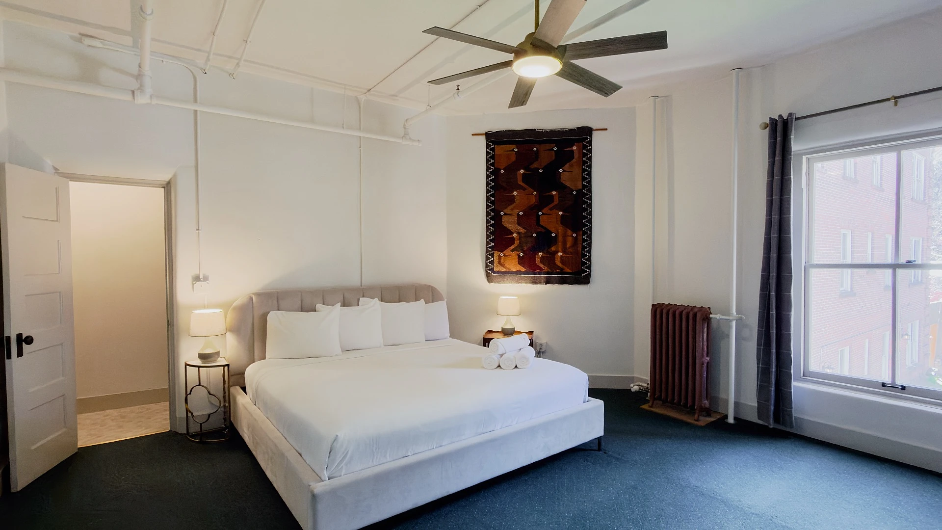 Bed with ceiling fan above in Room 218 at Hot Lake Springs Resort, open door to shower on left, window on right looking toward courtyard, dark blue/green carpet and open dark grey curtains.
