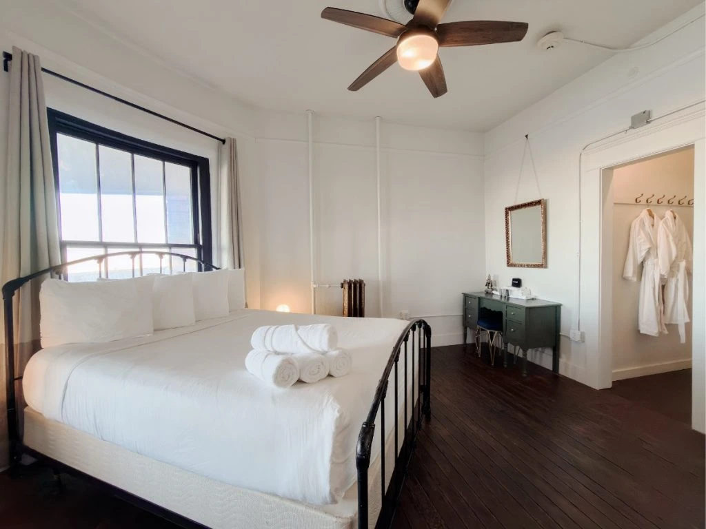 Bed with metal head and footboard in Room 223 at Hot Lake Springs Resort, window behind bed, olive green desk with mirror in corner, open doorway with two bathrobes hanging on right.