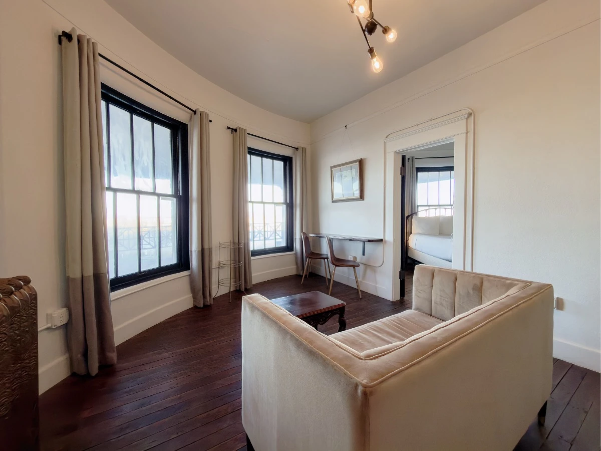 Curved exterior wall in Room 223 at Hot Lake Springs Resort with two windows, bar table and chairs after the windows, doorway to bedroom with partial bed view, sofa and square coffee table in foreground.