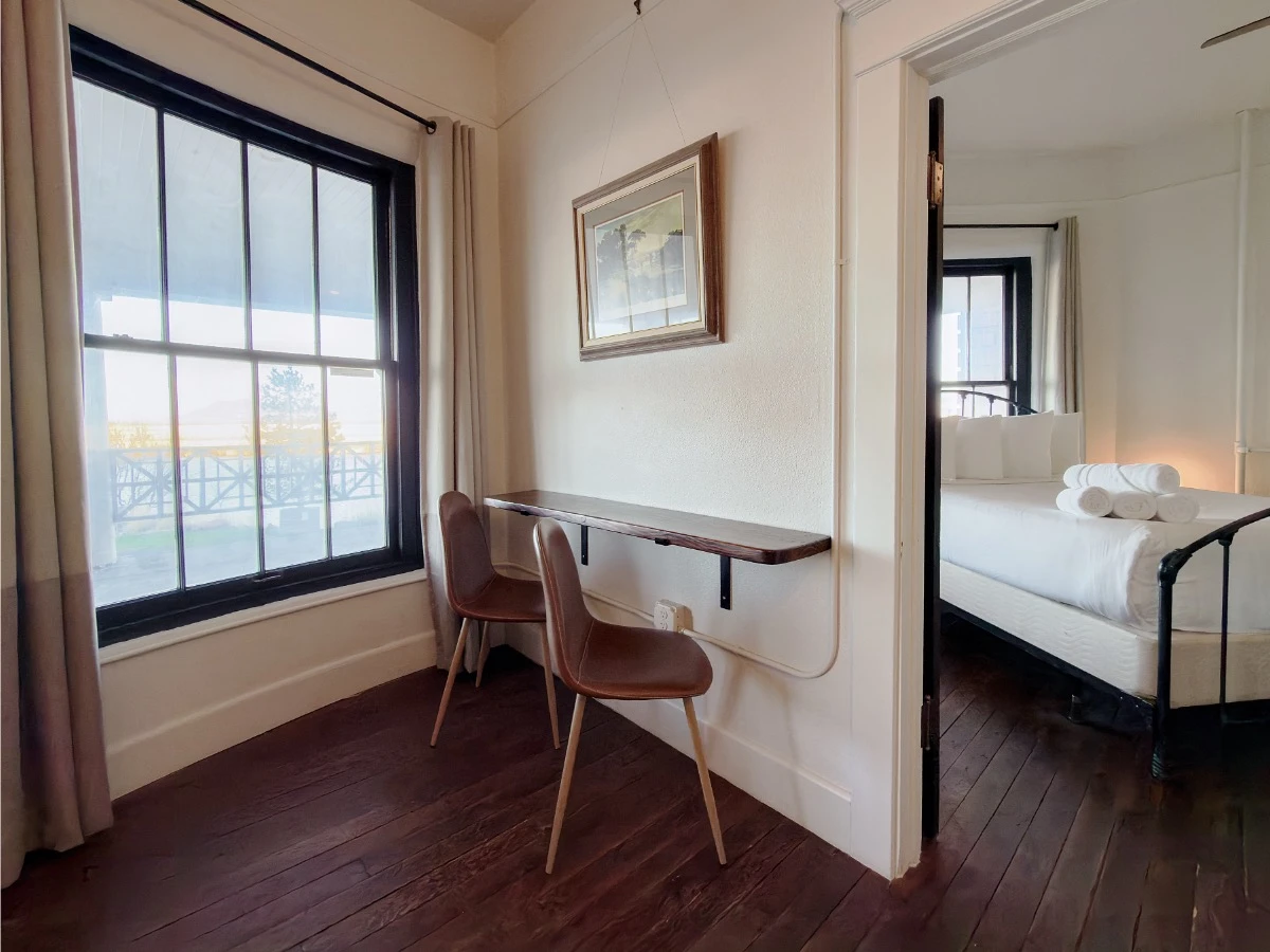 Living room in Room 223 at Hot Lake Springs Resort looking toward bedroom doorway, wall-mounted bar table with two chairs, window to left, partial view of bed through doorway.