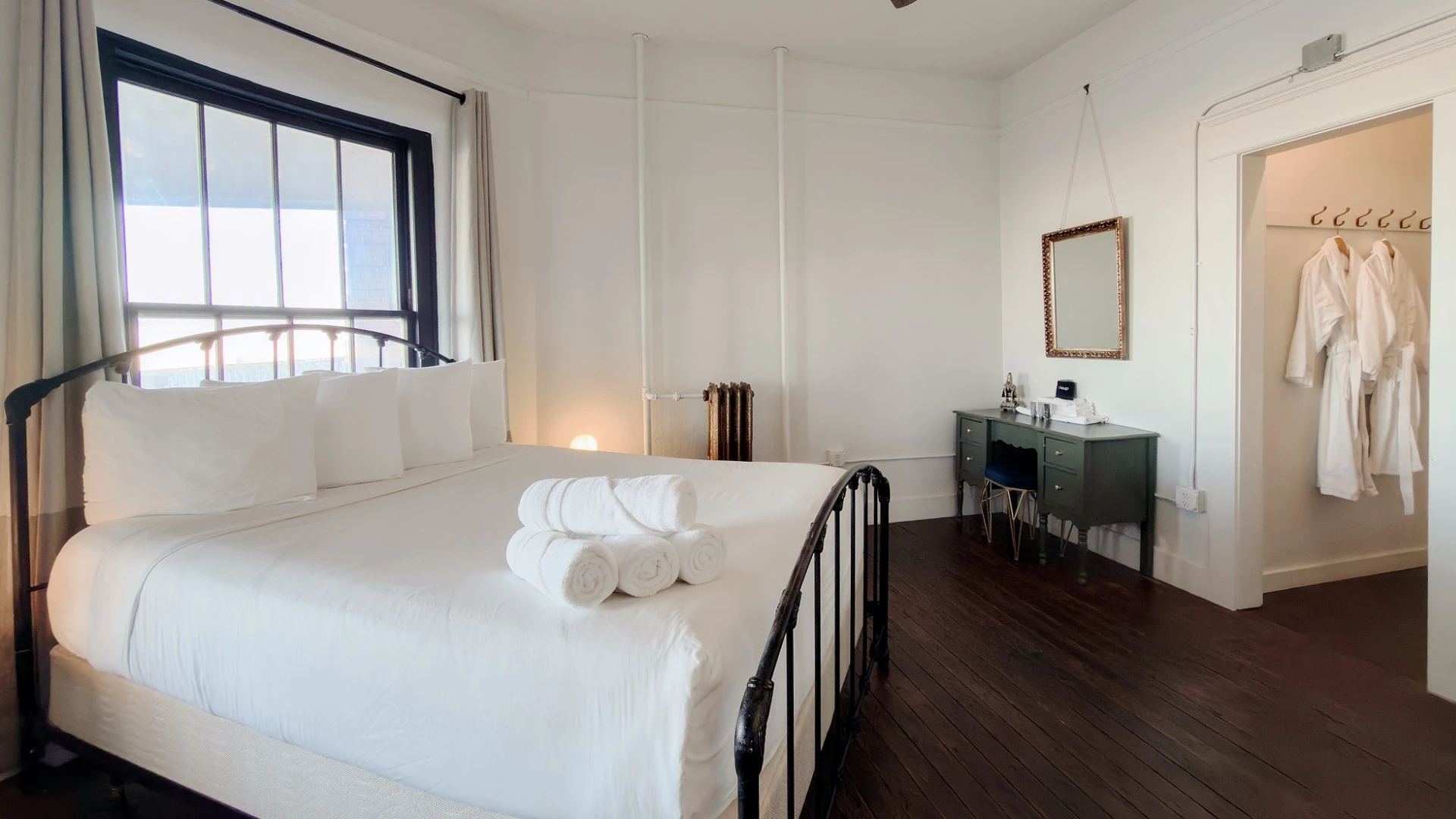 Bed with metal head and footboard in Room 223 at Hot Lake Springs Resort, window behind bed, olive green desk with mirror in corner, open doorway with two bathrobes hanging on right.