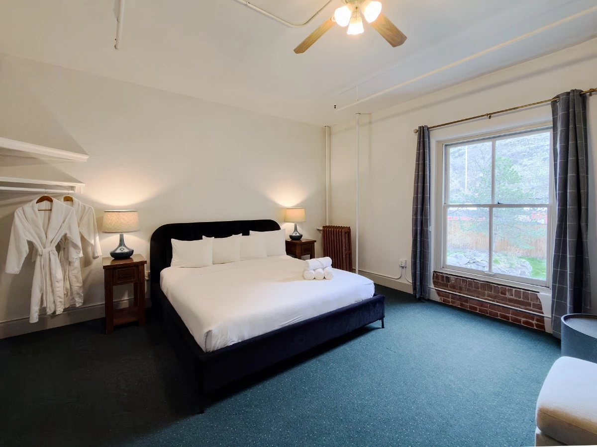Bed with two white bathrobes hanging on left in Room 224 at Hot Lake Springs Resort, window on right looking into courtyard below, dark blue/green carpet and open dark grey curtains.