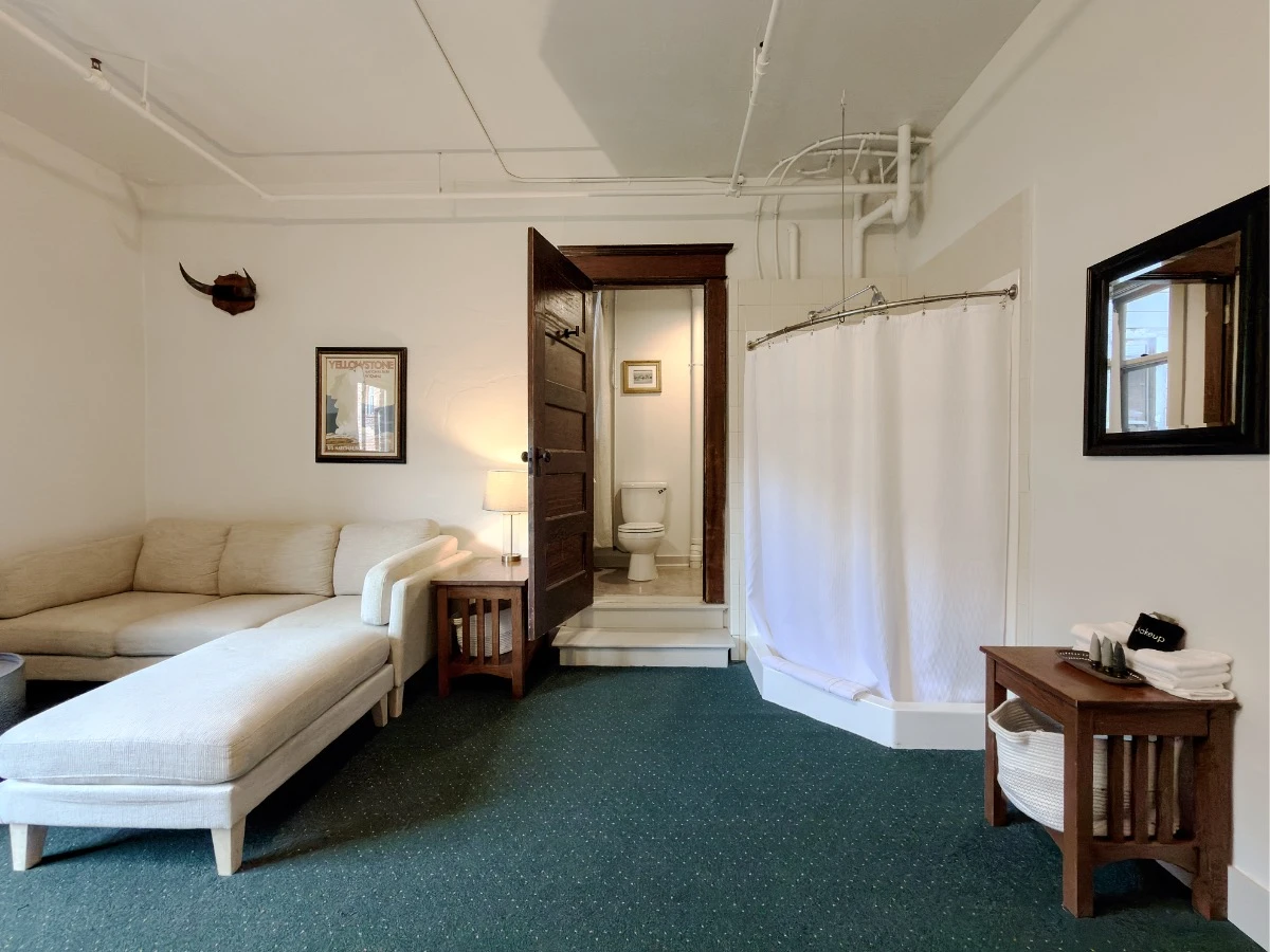 View toward bathroom in Room 224 at Hot Lake Springs Resort, dark wood door open into main room, off-white sofa with chaise lounge on left of door, corner stand-up shower with closed curtain on right of door, small wood end table with hand towels and mirror above on far right.