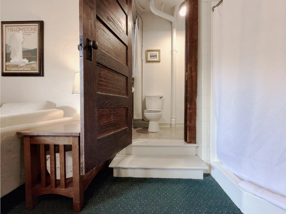 View into bathroom through open dark wood door in Room 224 at Hot Lake Springs Resort, bathroom is two steps up from main room, toilet visible in background, partial corner shower with closed curtain on right in main room.