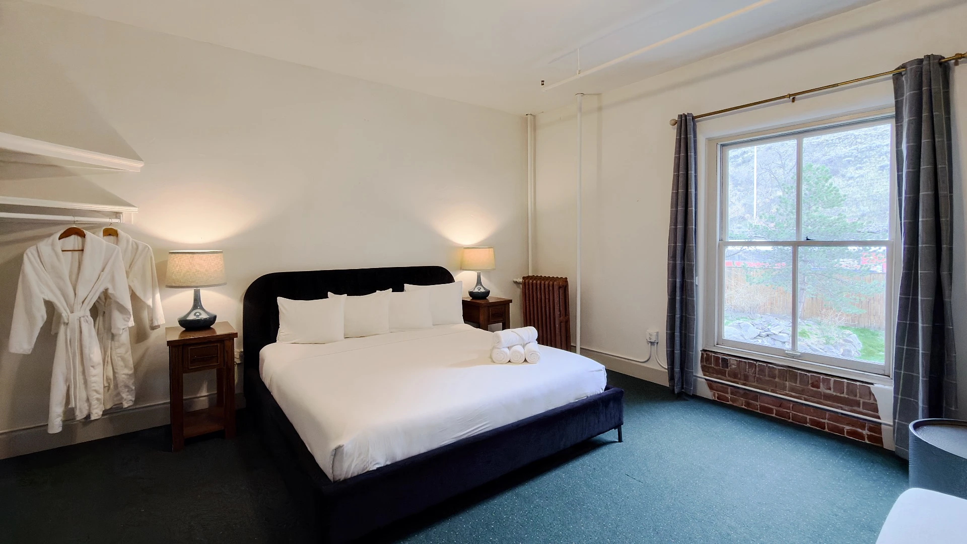 Bed with two white bathrobes hanging on left in Room 224 at Hot Lake Springs Resort, window on right looking into courtyard below, dark blue/green carpet and open dark grey curtains.