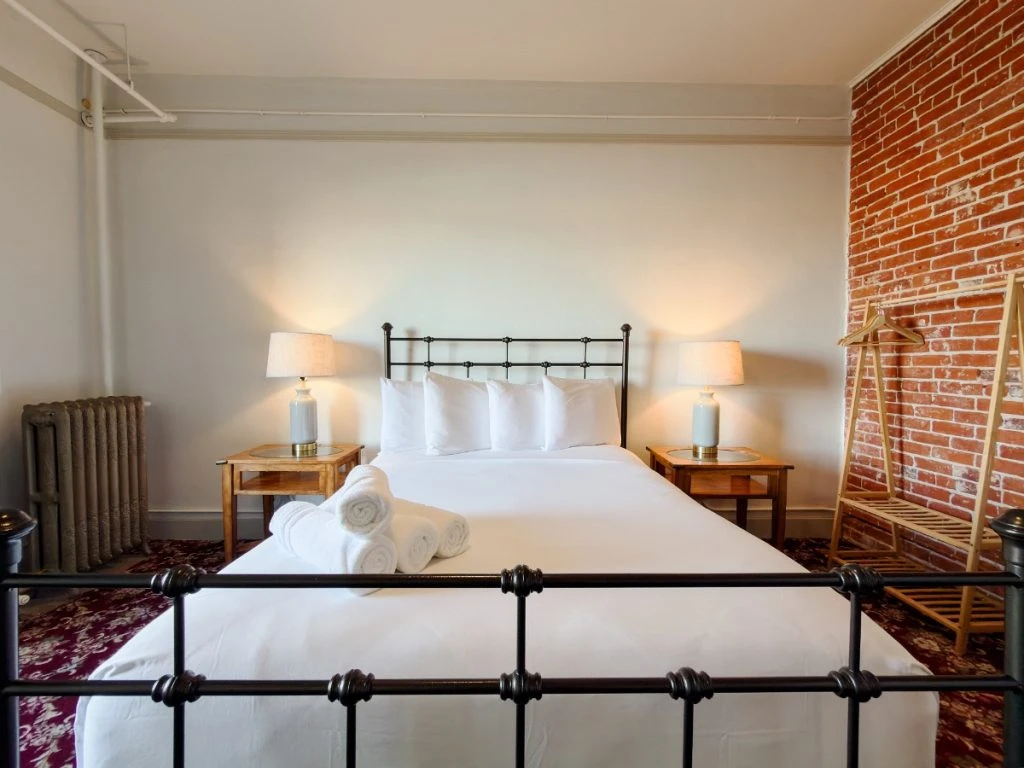 View from foot of bed in Room 227 at Hot Lake Springs Resort looking toward headboard with metal head and foot boards, exposed red brick on right wall, wood nightstands with lit lamps on each side.