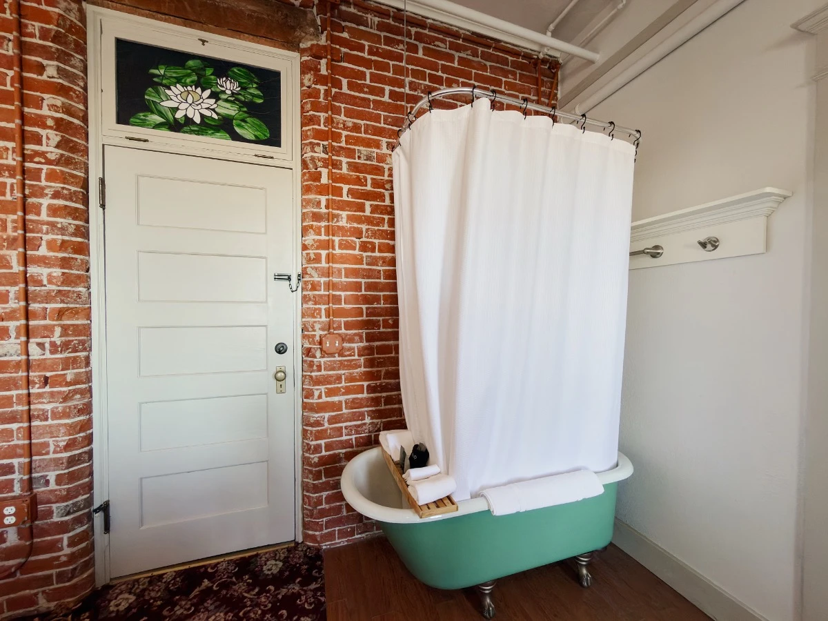 Clawfoot soaking tub in bedroom of Room 227 at Hot Lake Springs Resort against red brick wall, soaking tub shower curtain closed.