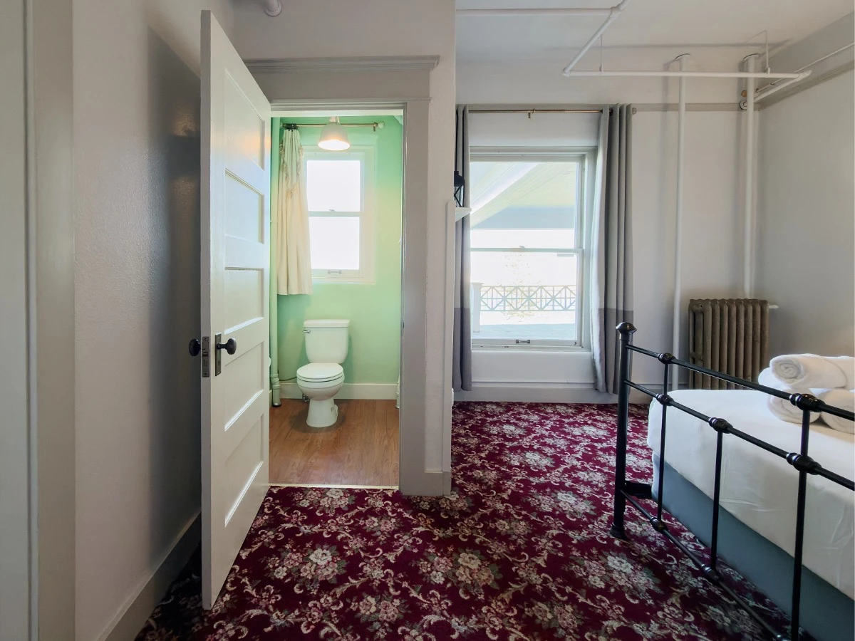 Bedroom in Room 227 at Hot Lake Springs Resort looking toward window and open bathroom door showing toilet, window visible to right of bathroom door.