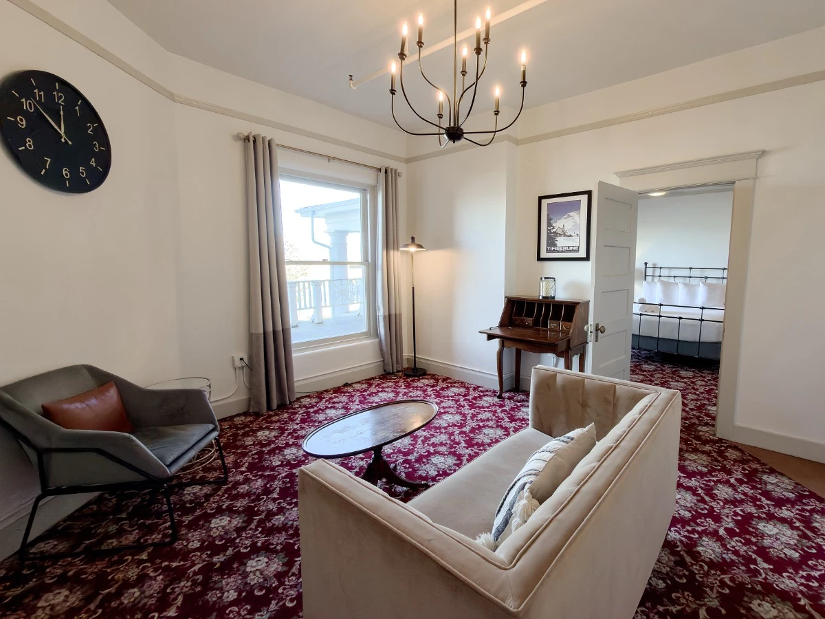 Living room in Room 227 at Hot Lake Springs Resort looking toward window and open bedroom door, loveseat sofa in foreground, wood writing desk to right of window, grey chair with large clock above on left.