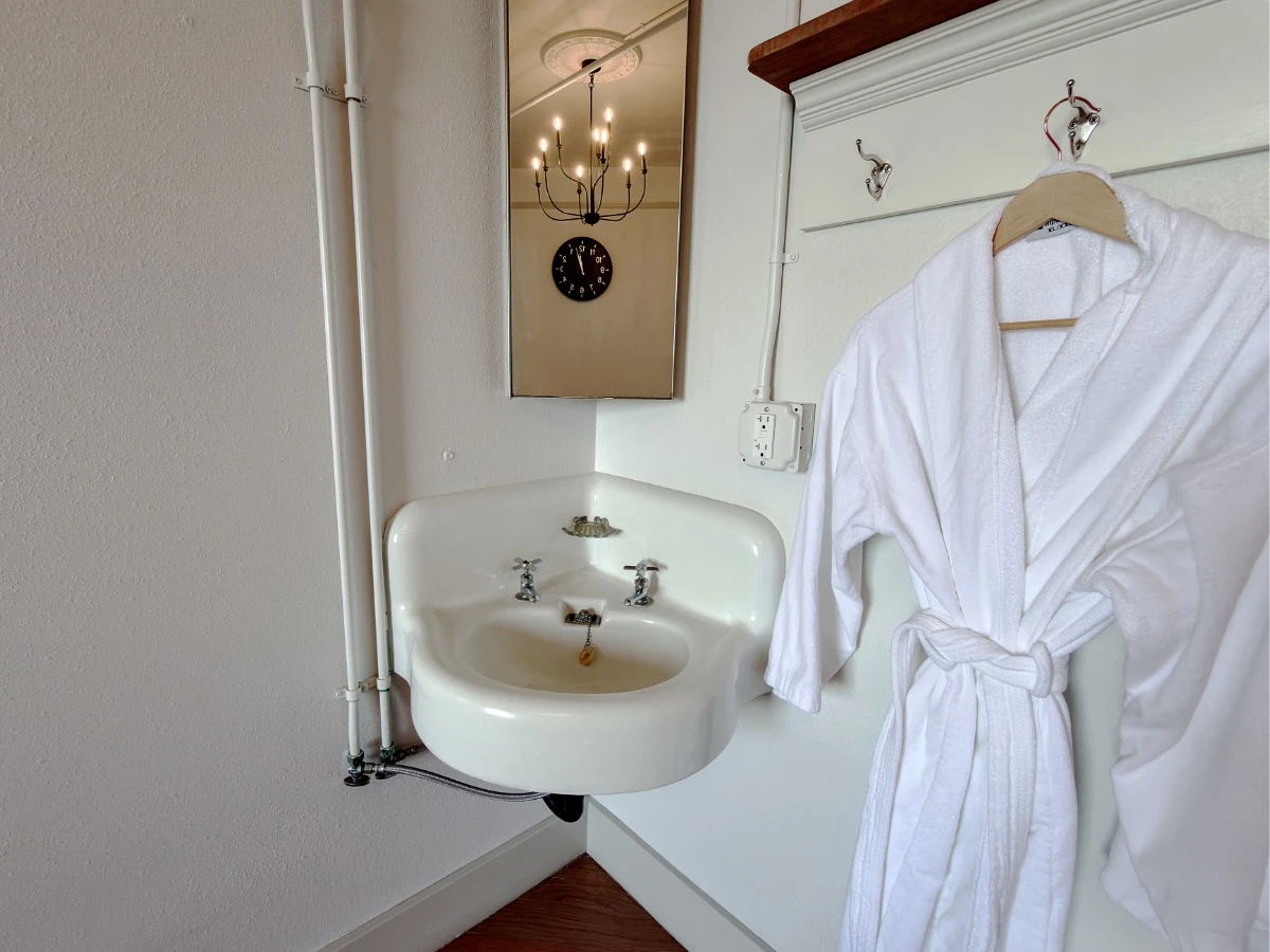 Corner sink with mirror in Room 227 at Hot Lake Springs Resort, clock and simple ceiling light reflected in mirror, white bathrobe hanging to the right of sink.