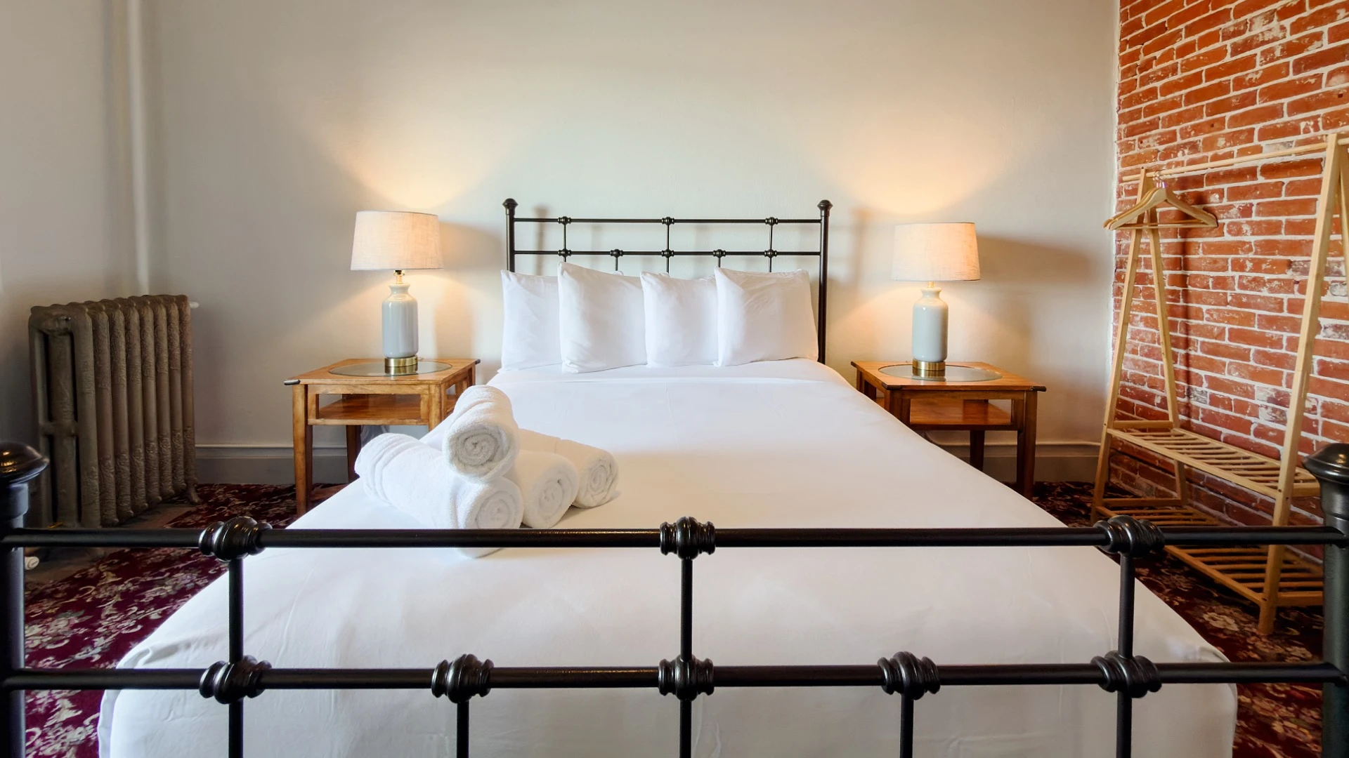 View from foot of bed in Room 227 at Hot Lake Springs Resort looking toward headboard with metal head and foot boards, exposed red brick on right wall, wood nightstands with lit lamps on each side.