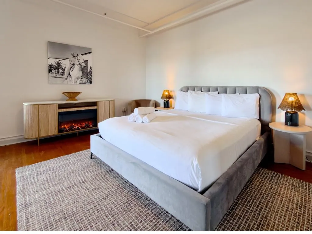 Master bedroom in Room 231 at Hot Lake Springs Resort with electric fireplace next to bed, black and white photo of man on horse above fireplace, wood floors with grey rug under foot of bed, sitting chair in back corner, nightstands with lit lamps on each side.