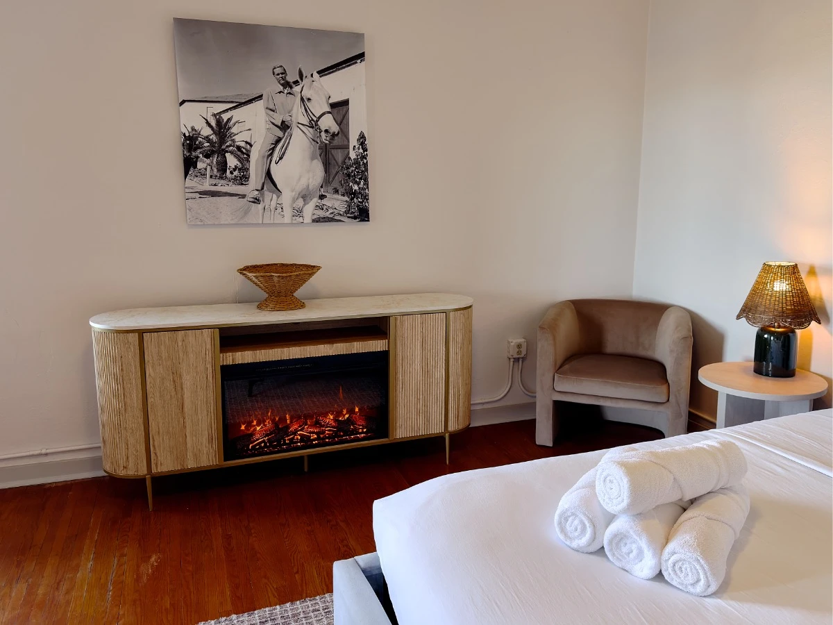 Master bedroom in Room 231 at Hot Lake Springs Resort looking across bed at electric fireplace, black and white photo of man on horse above fireplace, sitting chair to right of fireplace in corner.