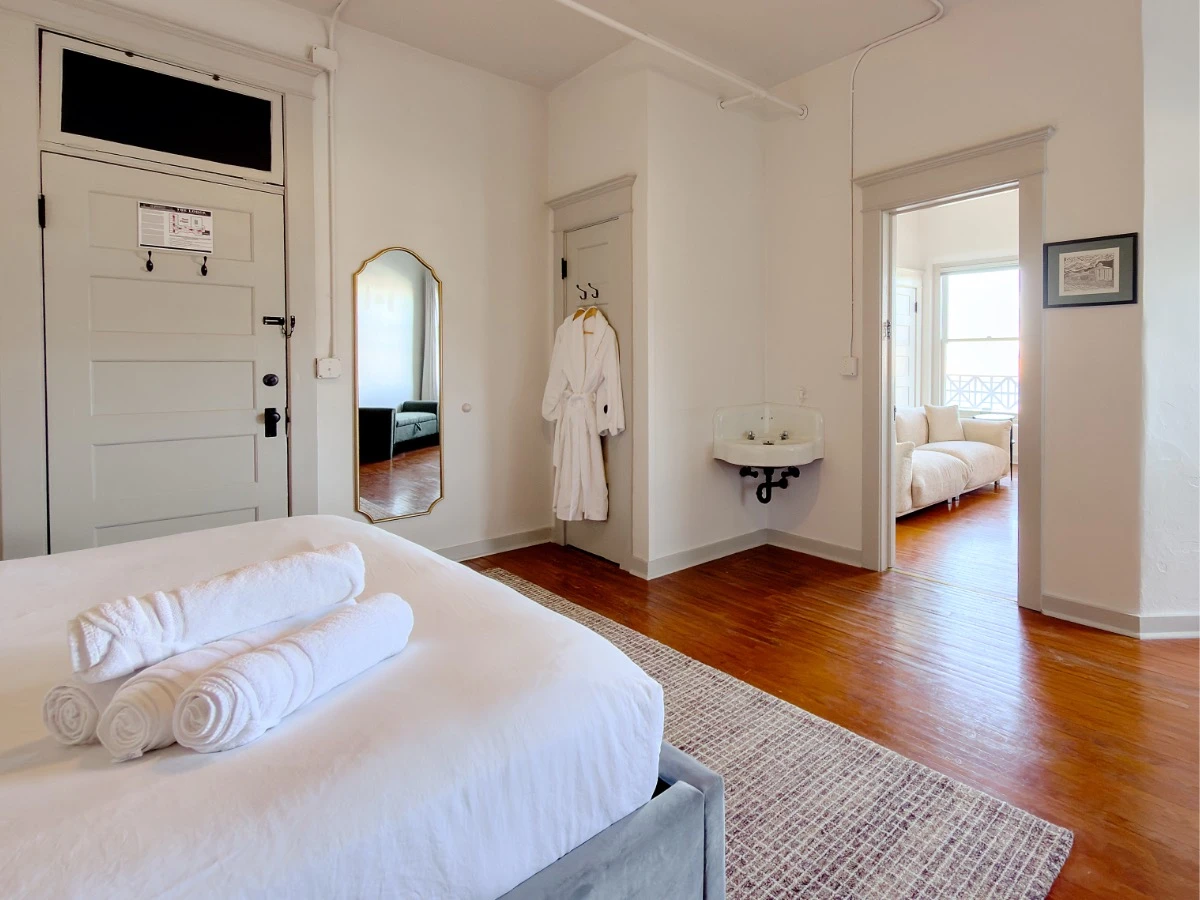 Master bedroom in Room 231 at Hot Lake Springs Resort looking across foot of bed toward living room, partial view of living room sofa visible, two white bathrobes hanging from closet door in corner, full body mirror on wall next to bathrobes.