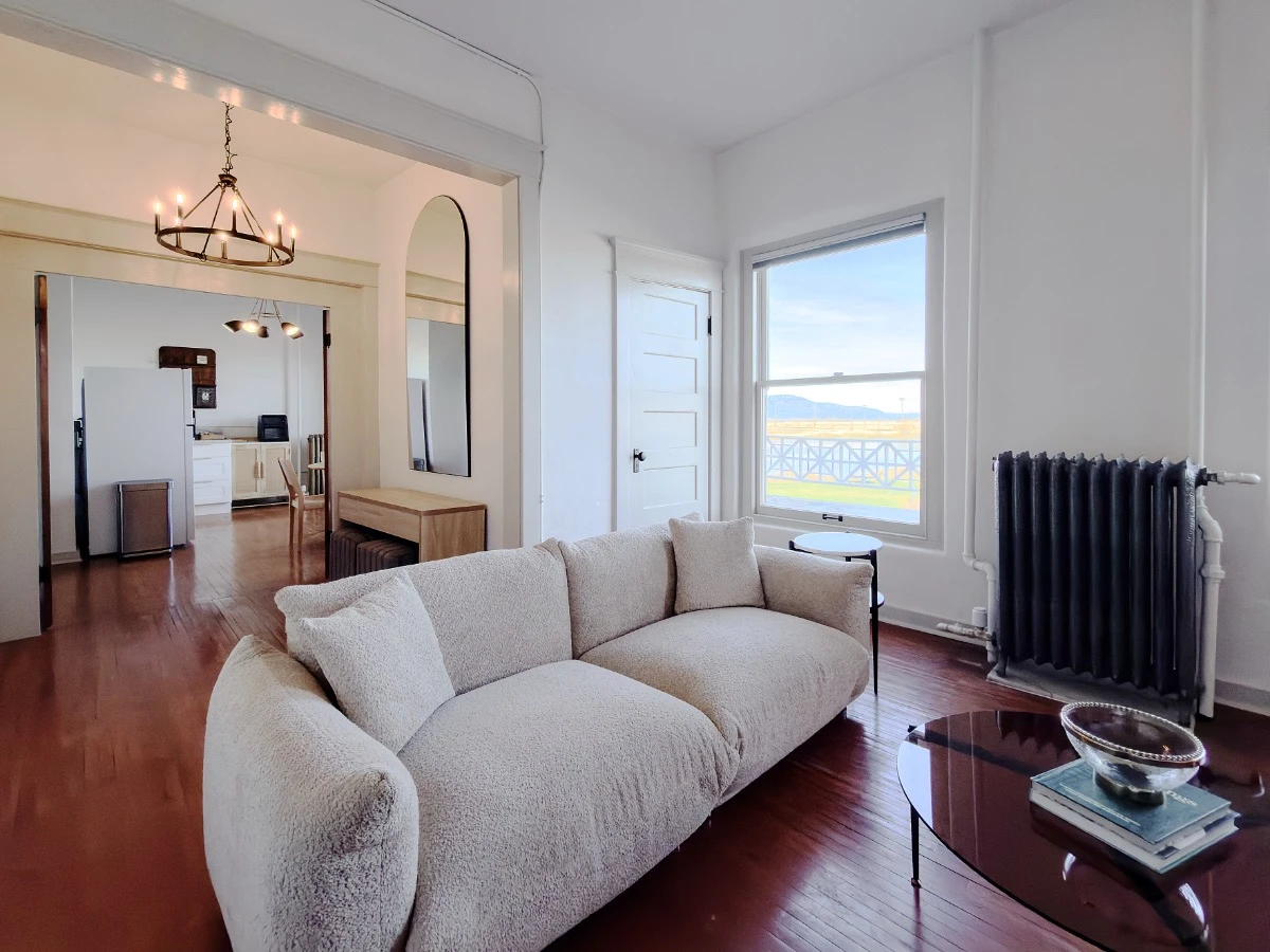Living room in Room 231 at Hot Lake Springs Resort looking toward kitchen, grey sofa in middle with coffee table in front, window with valley views in background, partial view of kitchen.